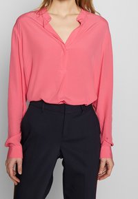 Lightweight, pink long-sleeve blouse with a V-neck and subtle ruffle detail at the collar; paired with dark tailored pants.