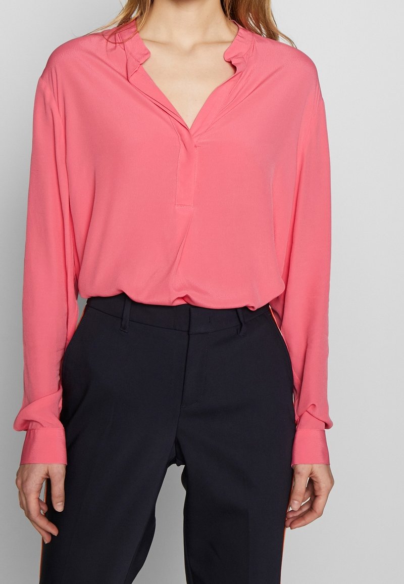 Lightweight, pink long-sleeve blouse with a V-neck and subtle ruffle detail at the collar; paired with dark tailored pants.