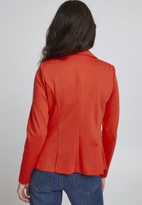Rode blazer met een getailleerde pasvorm, lange mouwen en een geknipte kraag. Glad materiaal met een schone achterkant, gecombineerd met blauwe jeans.