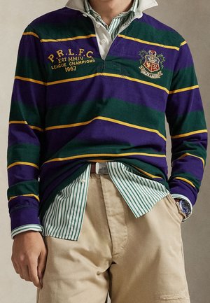 Homme portant un maillot de rugby rayé vert et violet sur une chemise à rayures vertes à boutons et un pantalon beige, avec montre et ceinture visibles.