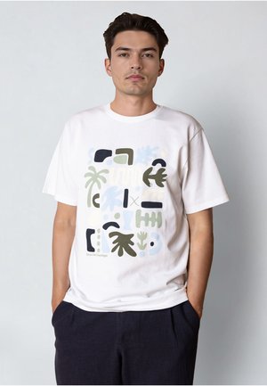 Junger Mann trägt ein weißes T-Shirt mit abstrakten grünen, blauen und schwarzen Formen und dunklen Hosen, steht vor einem schlichten hellen Hintergrund.