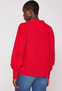 Blusa rossa realizzata in tessuto testurizzato con maniche a sbuffo e dettagli in pizzo ai polsini. Presenta un collo rotondo e una vestibilità rilassata.