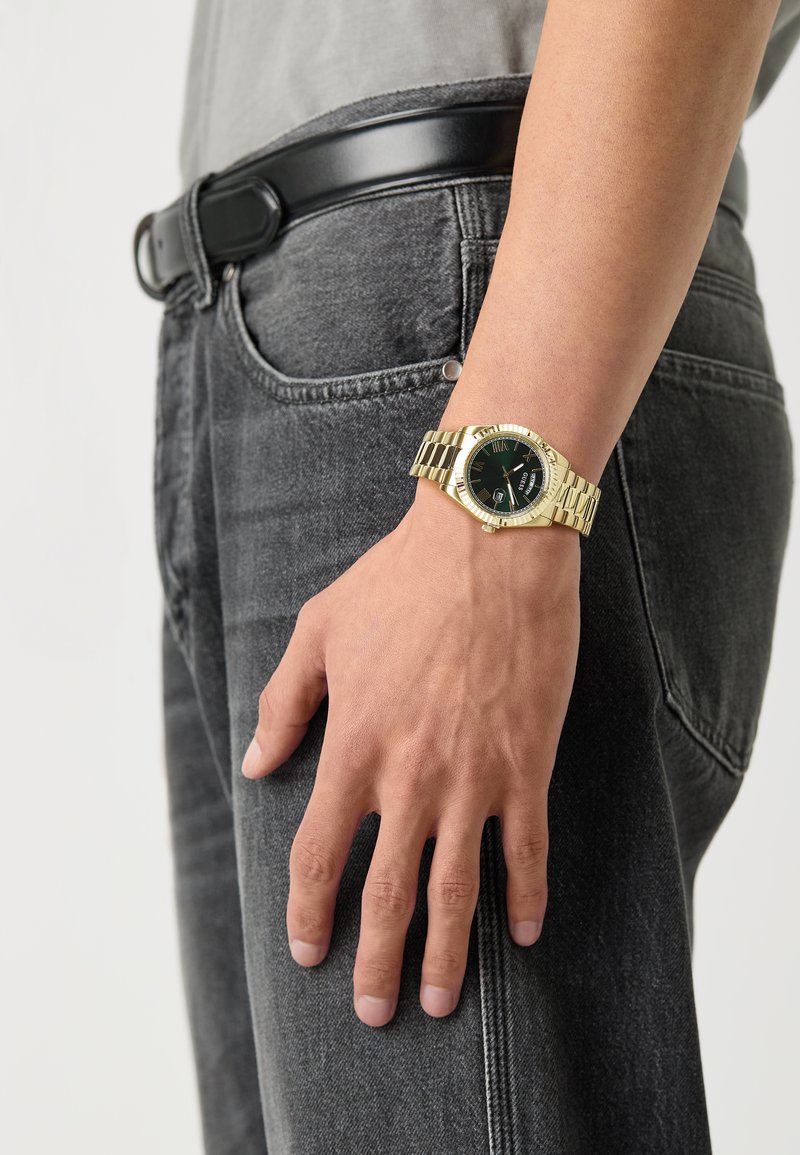 Montre en or avec un cadran noir et une fonction date, fixée à un bracelet en or. Un jeans en denim gris et une ceinture noire sont visibles.