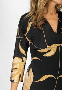 Musta mekko pitkillä hihoilla, jossa on suuria kultaisia kukkakuoseja ja V-pääntie. Sileä kangas, jossa on valuva tekstuuri.