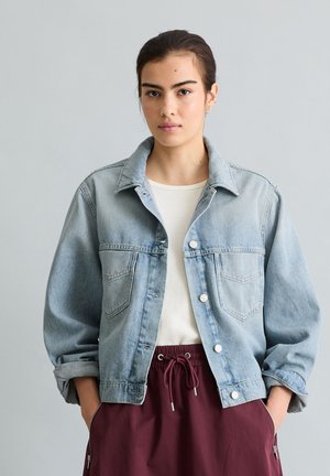 Junge Frau mit dunklen Haaren, die eine hellblaue Jeansjacke über einem weißen Top und burgunderrote Kordelzughosen trägt, steht mit den Händen in den Taschen.