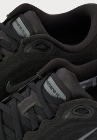 Svarta atletiska skor med texturerad meshovandel, vadderad krage och snören. Har en grå Nike-logotyp och branding på sidofliken.