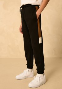 Pantalon de survêtement noir avec des accents marron et blanc, poches latérales, taille avec cordon de serrage et poignets côtelés, accompagné de baskets blanches.