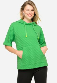 Hoodie verde trapuntato con maniche corte, cordino regolabile, tasca frontale e motivo testurizzato. Presenta maniche arrotolate e accenti luminosi.