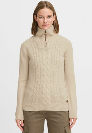 Beige kabelstrikket genser med høy krage, halv glidelås, ribbestrikkede mansjetter og nederkant. Myk tekstur; har et merke på venstre side.