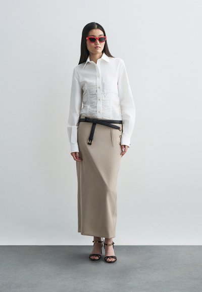 Camisa blanca con frontis texturizado, falda beige de talle alto, cinturón negro con hebilla de metal, gafas de sol rojas y tacones negros con tiras.