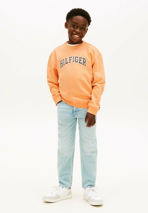 Smiling boy standing casually, wearing an orange "Hilfiger" sweatshirt, light blue jeans, and white sneakers on a plain white background.