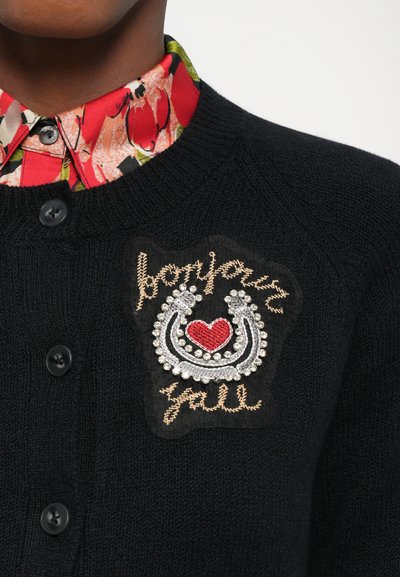 Black knit cardigan featuring a decorative patch with embroidery and rhinestones. Floral shirt collar visible underneath. Four buttons on front.