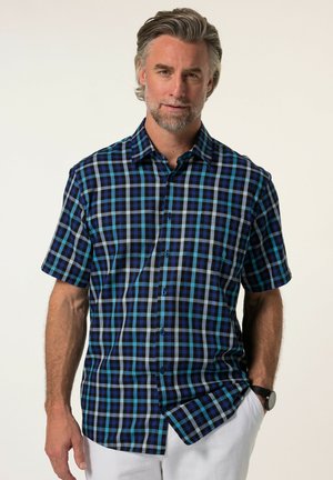 Middelbare man met grijs haar die een blauw-zwart geruit kort mouw overhemd en witte broek draagt tegen een lichte achtergrond.