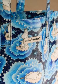 Niebieska torba na ramię z tkaniny z postaciami Moominów na ciemnym, gwiaździstym tle, z wzorami chmur i etykietą z napisem "Cath Kidston x Moomin."