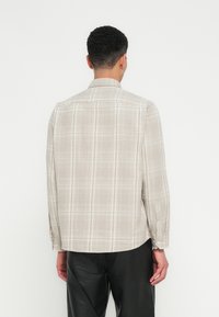 Hombre de espaldas vistiendo una camisa de cuadros beige de manga larga y pantalones negros frente a un fondo blanco liso.