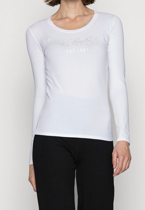 Femme portant un haut blanc ajusté à manches longues avec le texte "GUESS EST. 1981", associé à un pantalon côtelé noir.