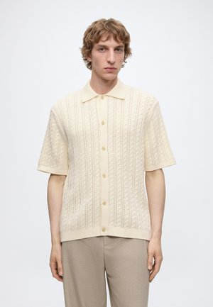 Jeune homme aux cheveux courts et bouclés portant une chemise en maille crème à manches courtes boutonnée et un pantalon beige sur un fond clair uni.