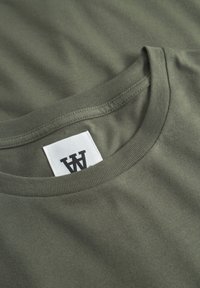 T-shirt col rond vert olive avec une étiquette rectangulaire blanche portant un logo "W" stylisé en noir, cousue à l'intérieur du col.