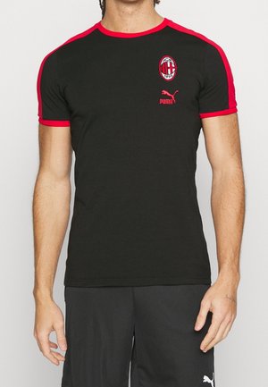 Homme portant un t-shirt noir AC Milan Puma avec des bordures rouges aux manches et au col, debout devant un fond clair uni.