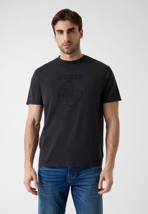 Homme portant un t-shirt noir Guess avec logo en relief et un jean bleu, debout devant un fond clair uni, face à l’avant.
