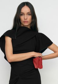 Fekete ruha szoborszerű vállrészlettel és texturált anyaggal, kis piros táskával párosítva, amely kontrasztot mutat a színben és az alakzatban.