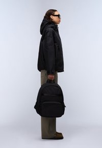 Svart ryggsäck tillverkad av texturerat tyg, med en framficka, sidofickor i nät och ett vadderat handtag. Modellen har på sig en svart jacka och khakifärgade byxor.