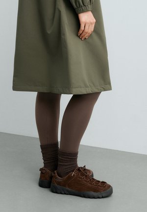 Legs wearing brown tights, brown knitted socks, and brown lace-up casual shoes standing on gray floor, olive green dress partially visible.