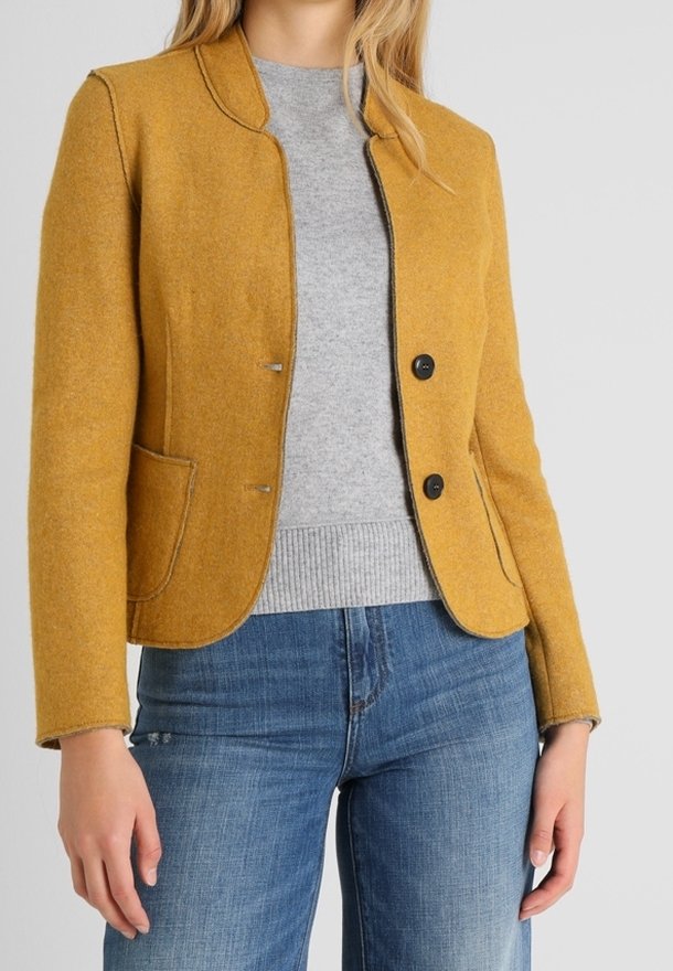 Femme portant un blazer boutonné jaune moutarde sur un pull gris clair, associé à un jean bleu, se tenant devant un fond uni.
