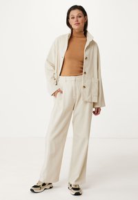 Béžová kordurová bunda s velkými kapsami, zkombinovaná s krémovým pleteným rolákem a volnými pruhovanými kalhotami. Tenisky mají černé akcenty.