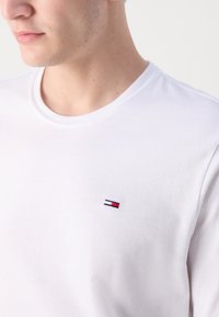 Camiseta blanca de algodón con un cuello redondo acanalado y un pequeño logo bordado en rojo, blanco y azul en la parte izquierda del pecho.