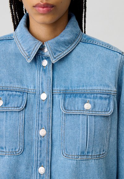 Giacca in denim blu chiaro con colletto a punta, bottoni a pressione metallici e due tasche sul petto con patta. Dettaglio di cucitura distintivo visibile.