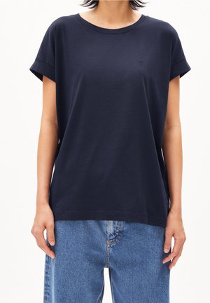 Person trägt ein schlichtes marineblaues Kurzarm-T-Shirt mit hochgekrempelten Ärmeln und hellblaue Jeans, gezeigt vom Hals bis zu den Hüften.