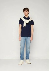 Fiatal férfi sötétkék pólót, világoskék farmert és fehér tornacipőt visel, fehér pulóver lóg a vállán, egy sima háttér előtt áll.