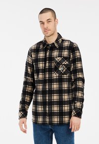 Camisa de franela a cuadros en negro y beige con un bolsillo en el pecho y etiqueta de la marca. Presenta un cierre frontal de botones y mangas largas.