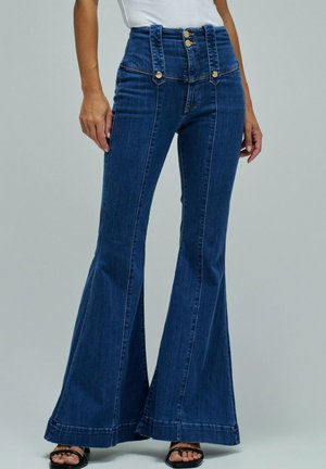 Femme portant un jean évasé taille haute bleu foncé avec des boutons dorés et des sandales noires à bout ouvert, debout devant un fond uni.