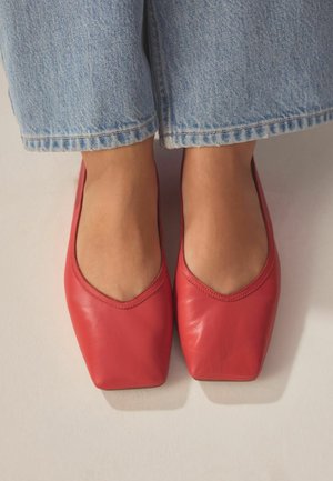 Stopy w czerwonych płaskich butach kwadratowymi noskami, zestawione z jasnoniebieskimi dżinsami na jasnej powierzchni.