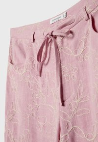 Pantalons en lin rose avec broderie florale blanche, un cordon de serrage à nouer à l'avant, et une étiquette de marque Stradivarius visible avec des détails de taille.