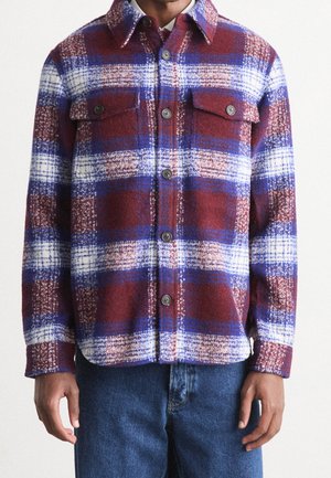 Mann trägt eine geknöpfte rote, blaue und weiße karierte Wollhemdjacke mit Brusttaschen, kombiniert mit blauen Jeans.