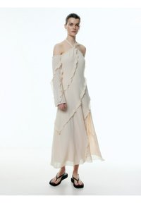 Jurkje van lichtbeige stof, met ruches, off-the-shoulder ontwerp, asymmetrische zoom en uitlopende mouwen. Zwarte slippers.