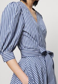 Blå och vit rutigt klänning med puffärmar, djup V-ringning, midjeband och sidofickor. Har en figurnära bodice och utsvängd kjol.