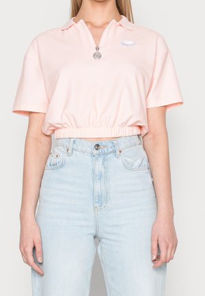 Woman wearing a light pink cropped Nike polo shirt with zipper and high-waisted light blue jeans against a plain background.