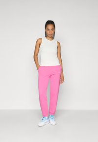 White sleeveless top and pink sweatpants, with elastic cuffs, logo detail, and white sneakers with blue accents, posed against a plain background.