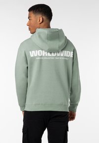 Grüne Kapuzenjacke aus weichem Stoff, mit einem großen weißen Grafikdruck auf der Rückseite, der den Text "WORLDWIDE" zeigt, und dezenten Rippenbündchen.