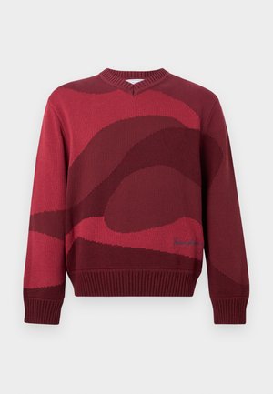 Κόκκινο πλεκτό πουλόβερ με V-λαιμόκοψη, με αφηρημένο κυματιστό μοτίβο σε πιο σκούρες αποχρώσεις του κόκκινου και ριμπ μανσέτες και τελείωμα. Μπλε κεντητή λεπτομέρεια στο κάτω μέρος.