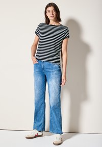 T-shirt noir et blanc rayé avec des manches retroussées, associé à un jean évasé bleu clair. Le modèle porte des baskets blanches avec des accents rouges.