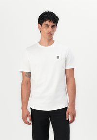 Weißes Baumwoll-T-Shirt mit Rundhalsausschnitt, kurzen Ärmeln und einem kleinen schwarzen Wappenlogo auf der linken Brust. Glatte Textur.