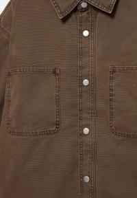 Brązowa koszula jeansowa z dwoma naszywanymi kieszeniami z przodu i srebrnymi zatrzaskami na środku, posiadająca kołnierzyk i widoczne przeszycia szwów.