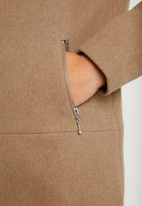 Hnědá tkanina ze směsi vlny s viditelnou kapsou na zip, která má stříbrnou tahací úchytku a hladkou strukturu. Minimalistický design s čistými liniemi.