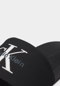 Gros plan d'une sandale noire à semelle plate avec une bride texturée ornée d'une broderie du logo Calvin Klein en blanc et gris.