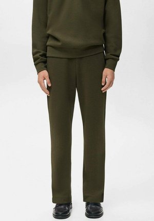 Hombre vistiendo pantalones rectos de color verde oliva, una camiseta de manga larga a juego y zapatos de cuero negros, de pie contra un fondo claro y liso.